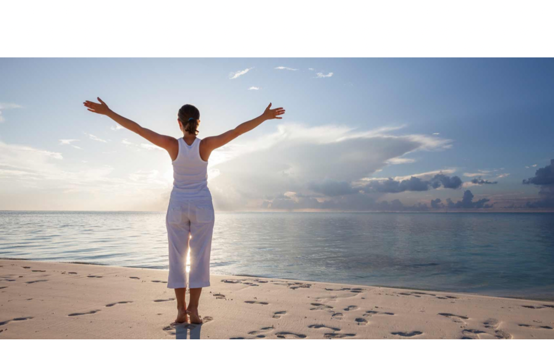 This is a picture of a young lady standing in the sunshine on a beach with her hands held outstretched. She is wearing a white t-shirt and white pants.