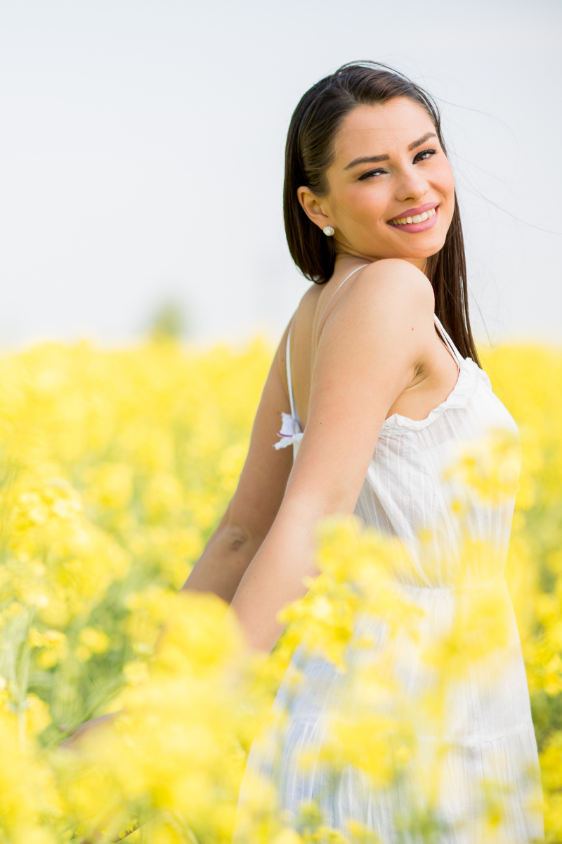 This an image of a girl with dark hair walking through a field of yellow flowers. She is looking directly at the camera. She is wearing a white dress. Looking back over her shoulder and smiling.