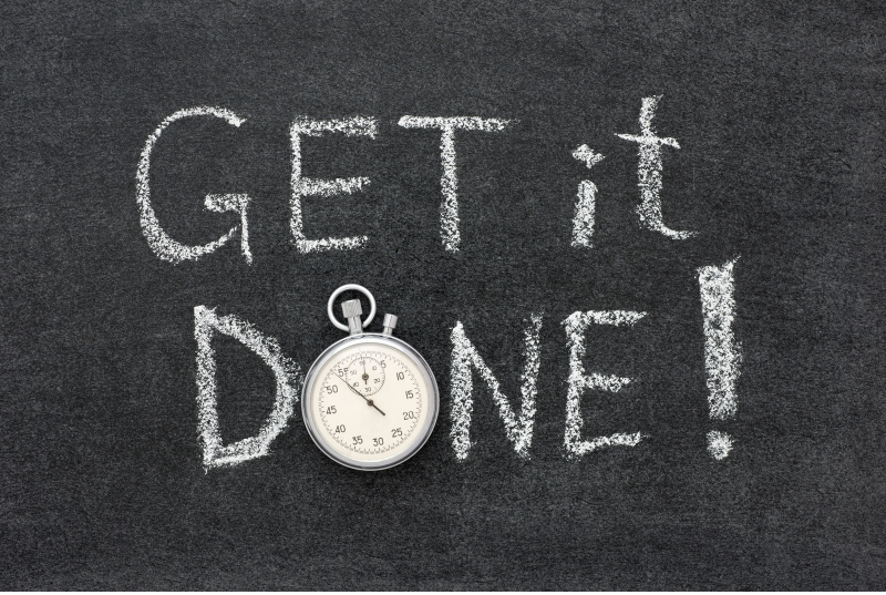 This is an image of text on a blackboard. The text it written with white chalk. The text says get it done. The O in done a has been replaced with a pocket stop watch.