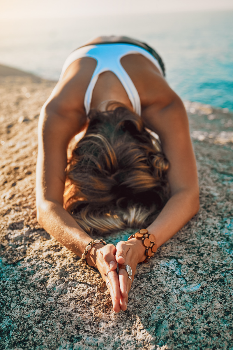 This is an image of a girls in a yoga pose on the beach. She is bent forward with her hands stretched in front of her. She is wearing a white top. She has bracelets on that are made of wood.